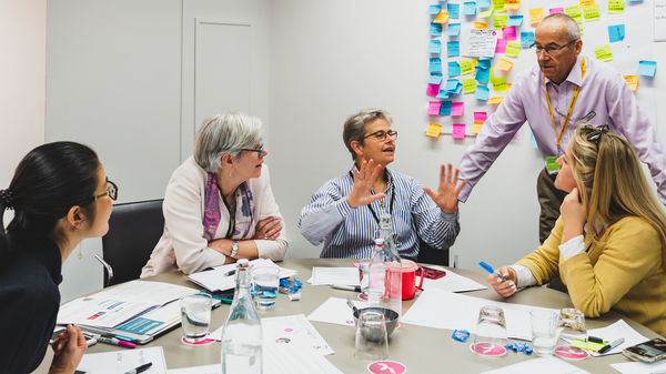 A group of six individuals around a conference table engaged in what appears to be a discussion or meeting. Their faces are obscured for privacy. The table is scattered with papers, water bottles, and pens, indicating an active work session. A whiteboard covered with colorful sticky notes is visible in the background, suggesting brainstorming or project planning activities. One individual at the head of the table is gesturing with their hands, possibly explaining a point or leading the conversation.